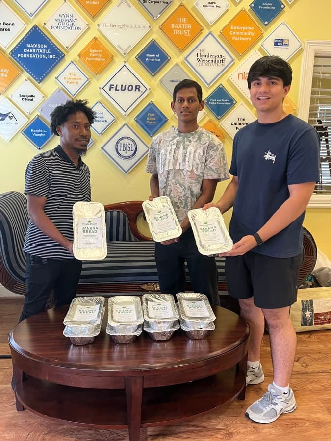 Sid Sharma and two volunteers holding labeled Waste2Taste banana bread loaves in a community center lobby